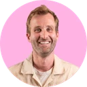 Homme souriant avec barbe légère, portant une chemise beige sur fond rose.