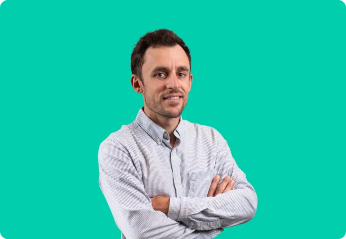 Homme souriant aux bras croisés portant une chemise claire sur fond turquoise uni.