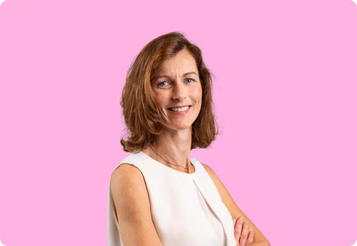 Femme souriante aux cheveux bruns mi-longs, portant un haut blanc, fond rose uni.