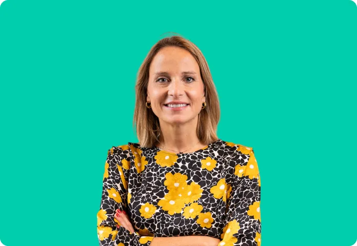 Femme souriante aux cheveux blonds avec un haut noir à fleurs jaunes, bras croisés sur fond turquoise.