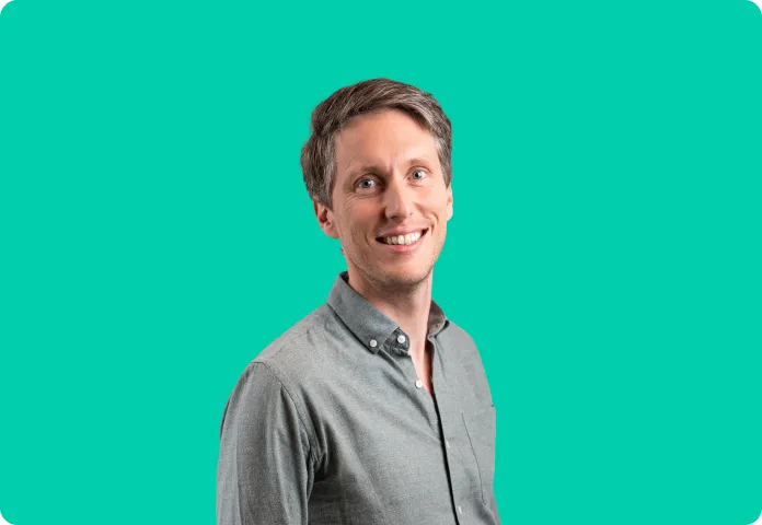 Homme souriant portant une chemise grise sur un fond turquoise uni.