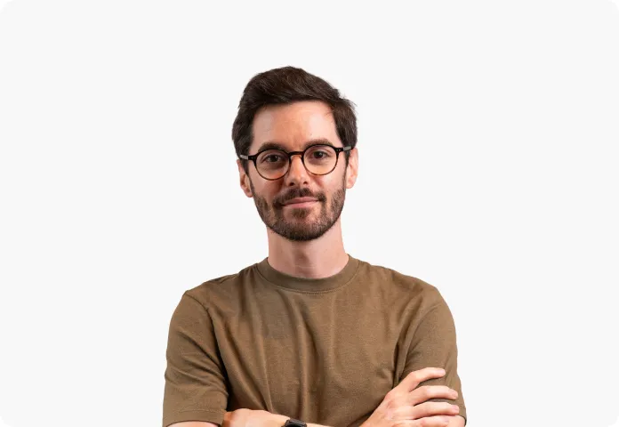 Homme souriant avec barbe, lunettes rondes, et t-shirt marron, bras croisés sur fond blanc.