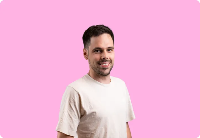 Un homme souriant avec un t-shirt beige sur fond rose uni.