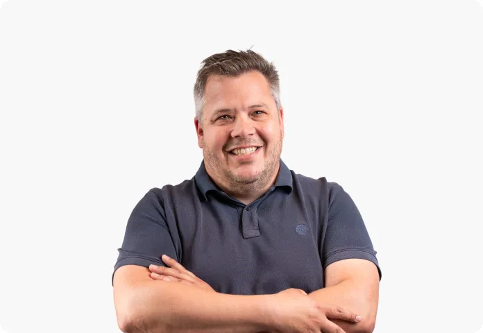 Homme souriant aux cheveux gris portant un polo bleu avec les bras croisés.