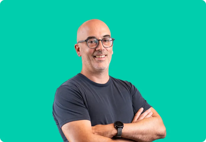 Homme chauve souriant avec lunettes et montre, bras croisés sur fond turquoise.