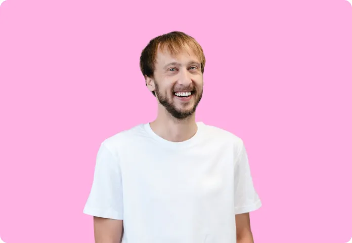 Un homme souriant avec barbe et t-shirt blanc devant un fond rose uni.