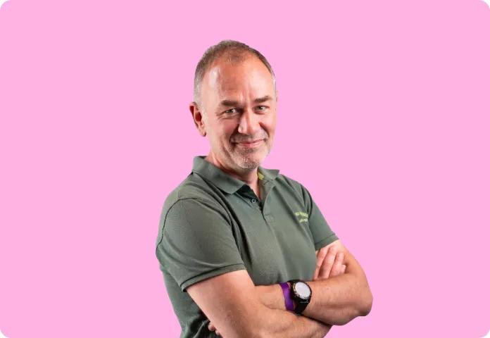 Homme souriant bras croisés portant un polo vert et une montre sur fond rose.