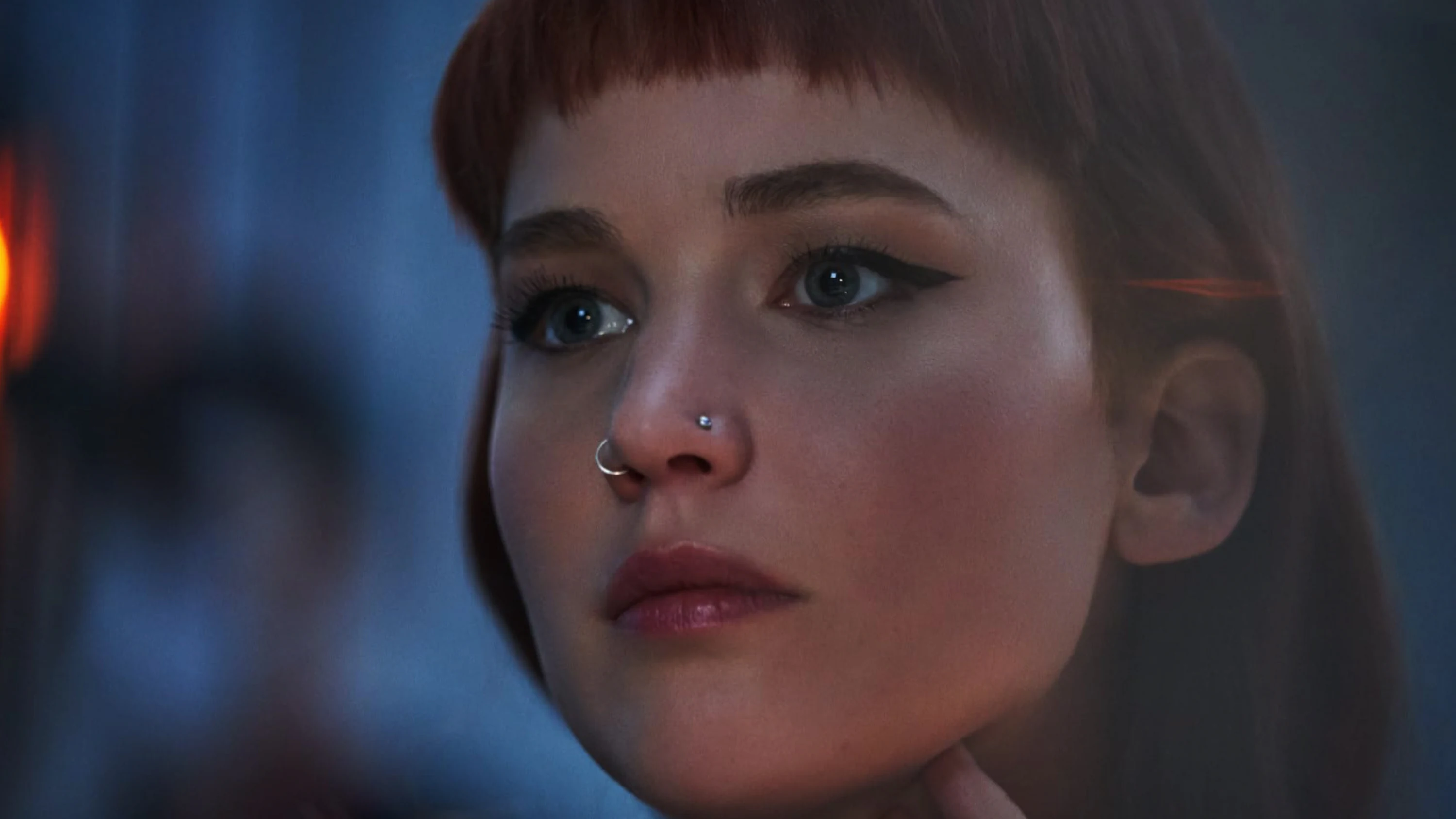 Close-up of a woman with short red hair, blue eyes, nose piercings, and subtle makeup looking thoughtfully.