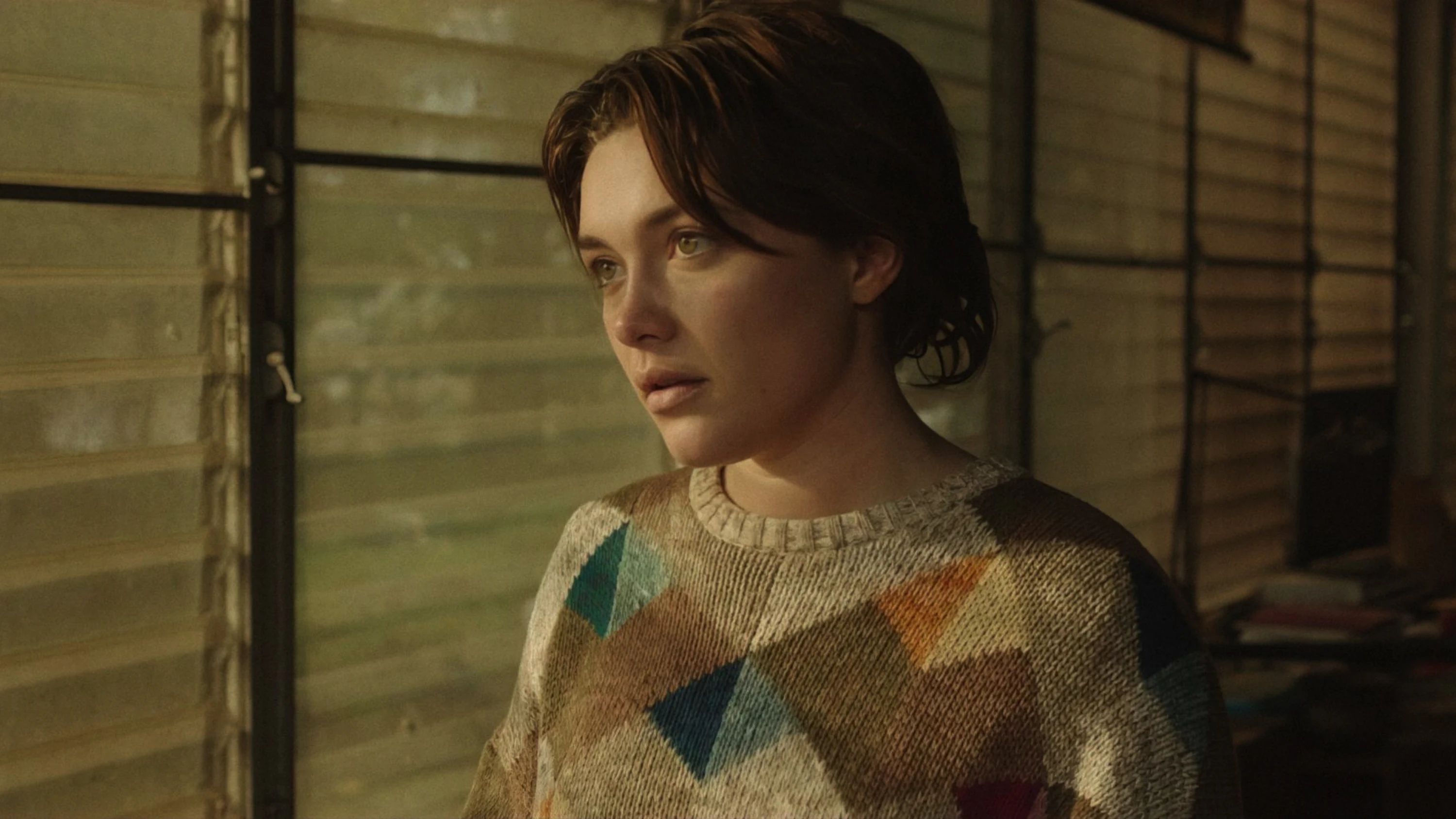 Woman with short brown hair wearing a patterned sweater looking pensively out a window.