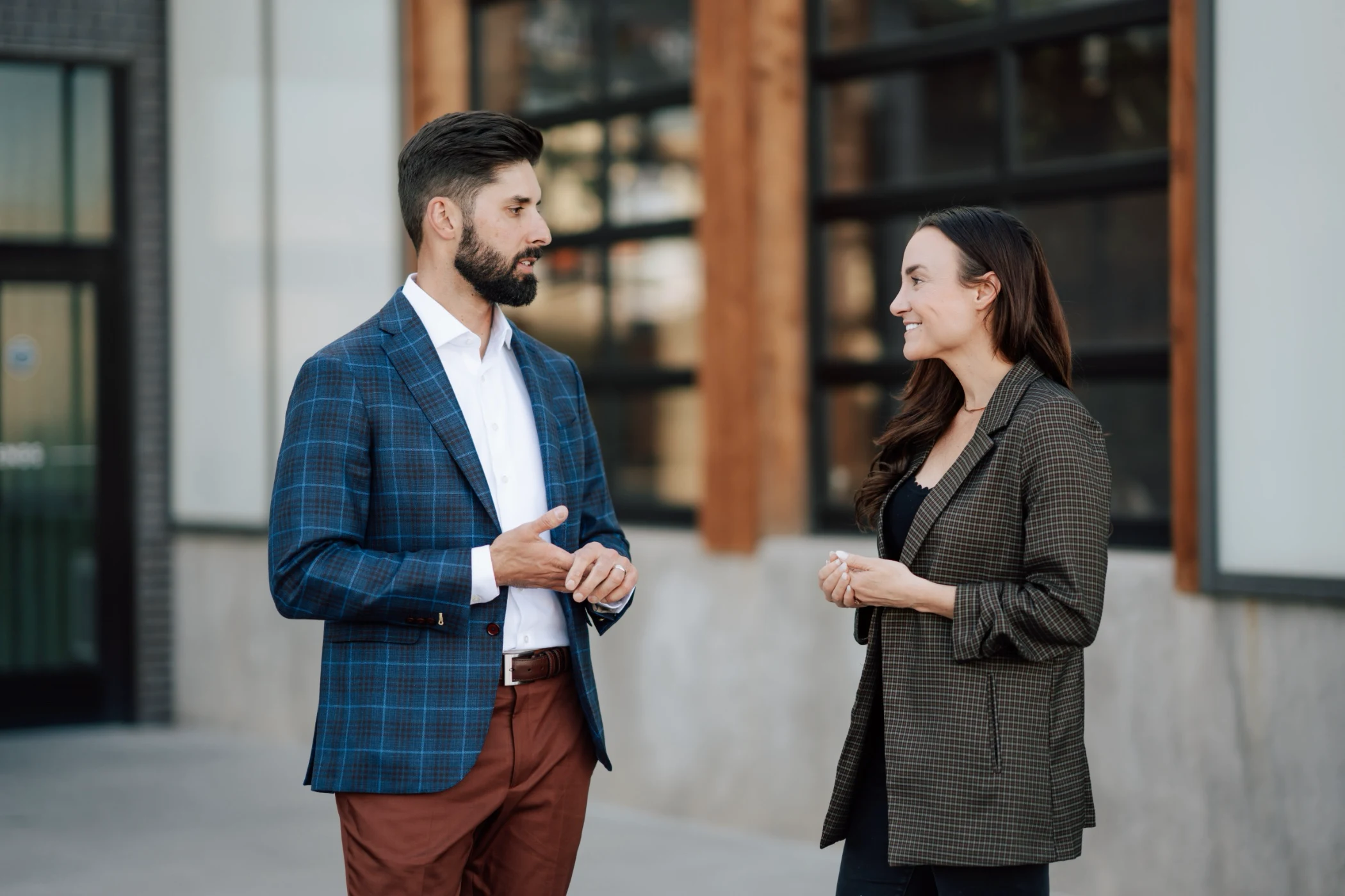 A man in a blue plaid blazer and brown pants talking to a woman in a checked blazer standing outdoors near a building.