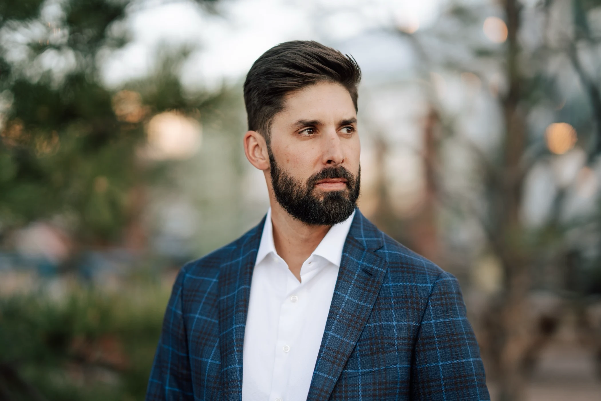 Bearded man in a blue checkered blazer and white shirt looking to the side with an outdoor blurred background.
