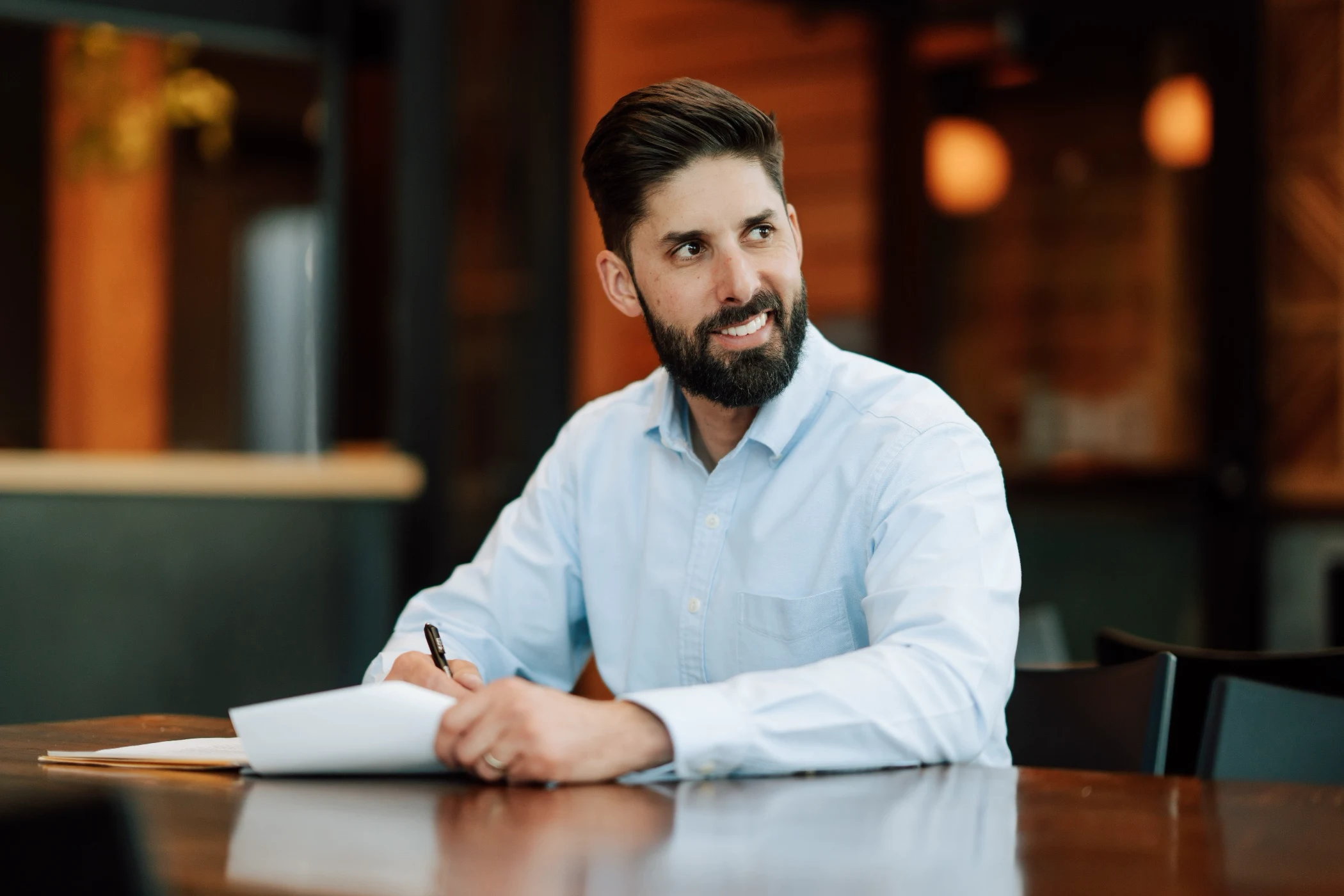 Smiling man with a beard in a light blue shirt sitting at a table holding a pen and paper.