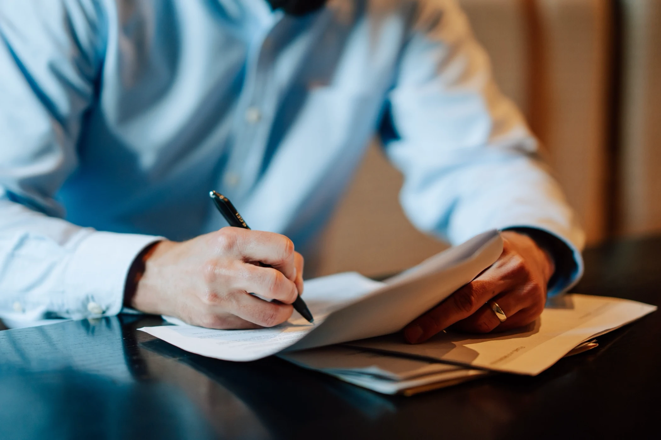 Person in a light blue shirt writing on documents with a pen on a dark table.
