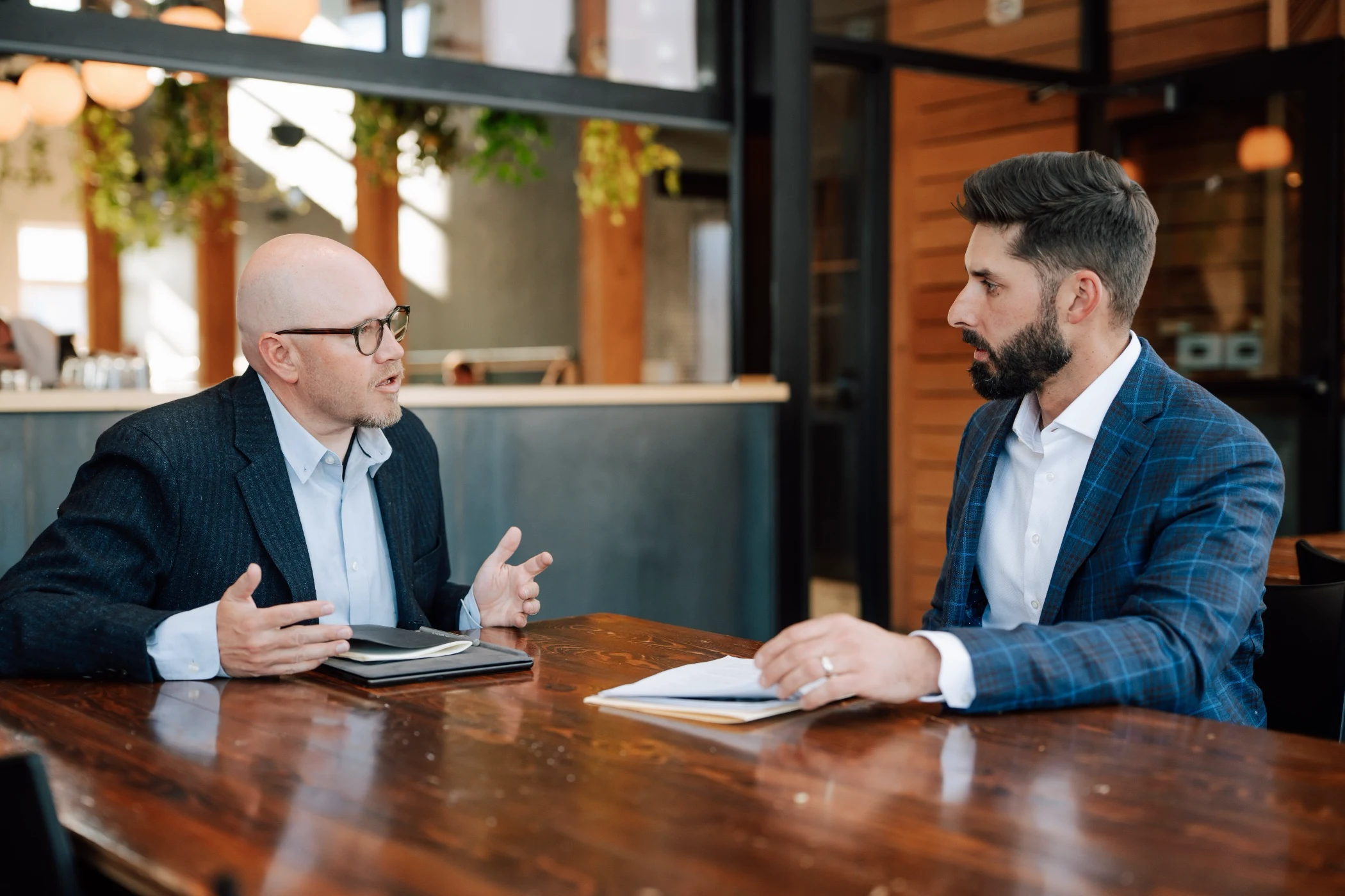 Two men in business attire having a serious discussion at a wooden table in a modern office setting.