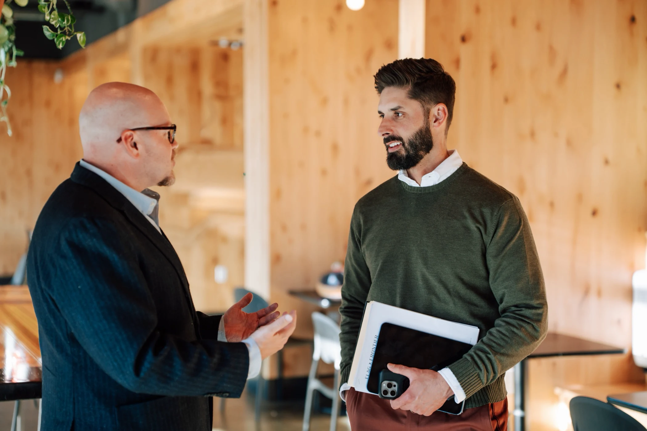 Two men conversing indoors, one bald man in a suit gesturing while the bearded man in a green sweater holds a tablet and phone.