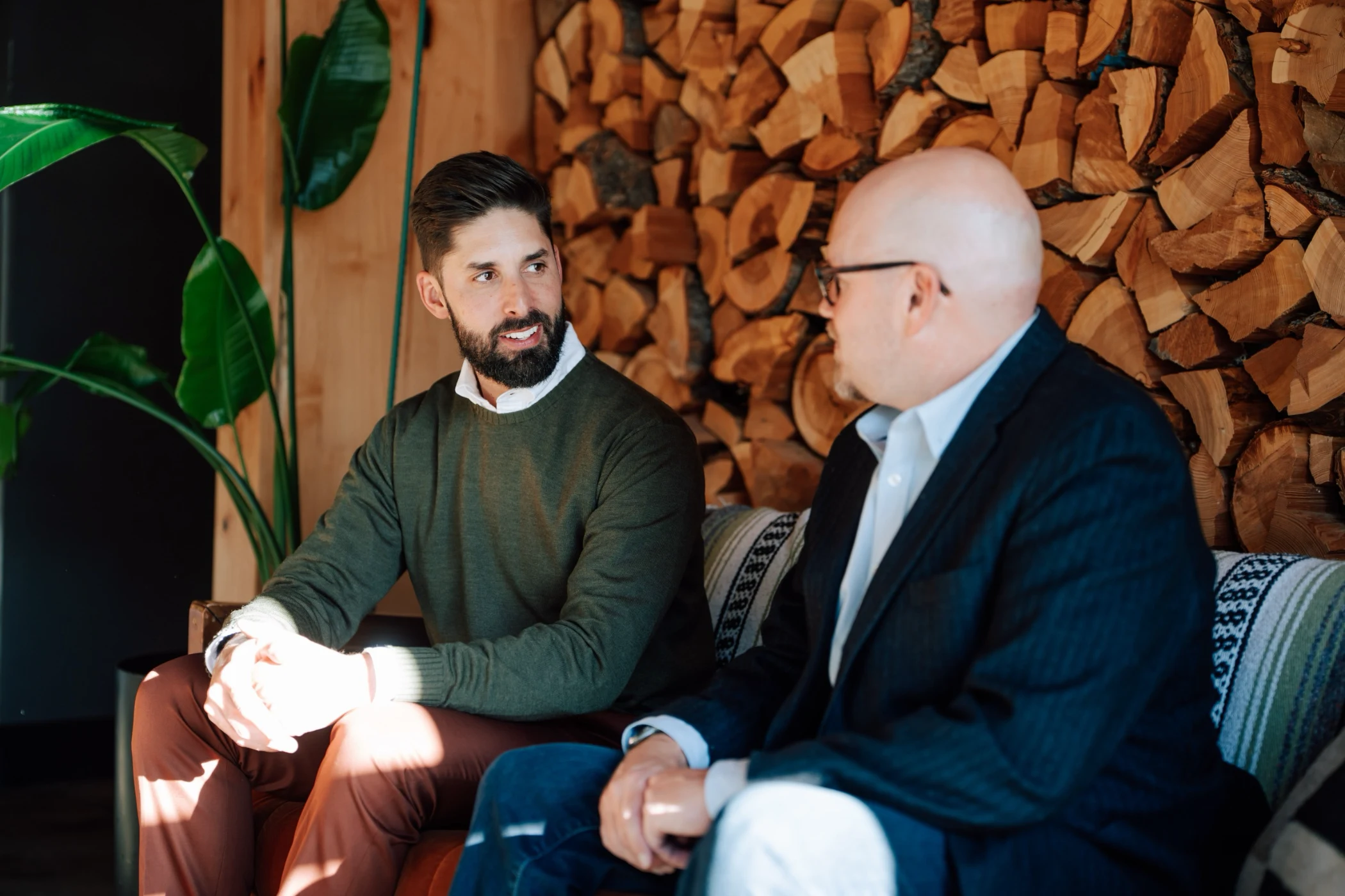Two men sitting indoors on a couch, engaged in conversation, with a wood log wall and green plant behind them.