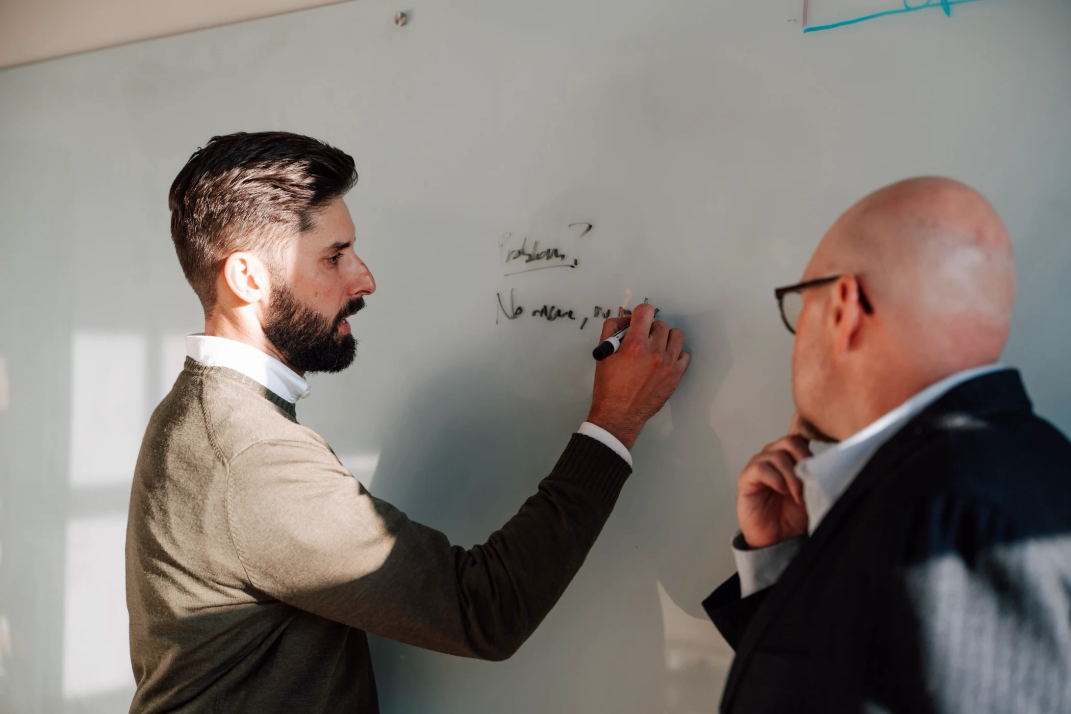 Man with beard writing on whiteboard while another bald man in glasses observes thoughtfully.