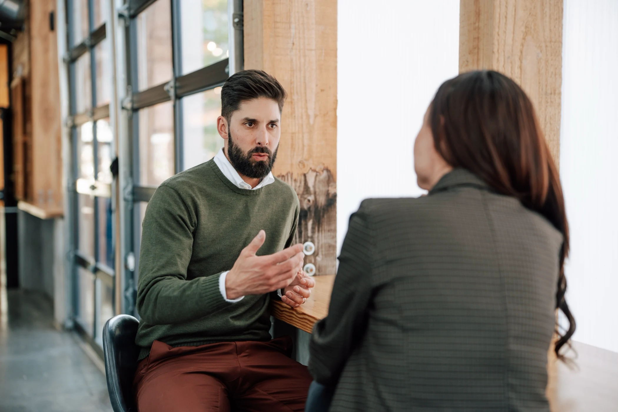 Man in green sweater gesturing while talking to a woman in a plaid jacket at a wooden counter by a window in a modern indoor setting.