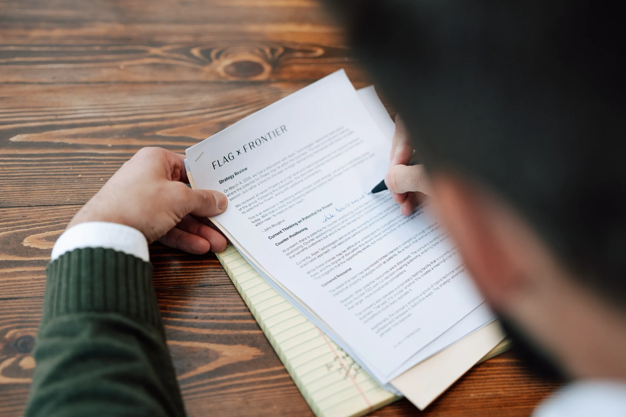 Person holding a document titled 'FLAG x FRONTIER' and writing notes with a pen over a wooden table.