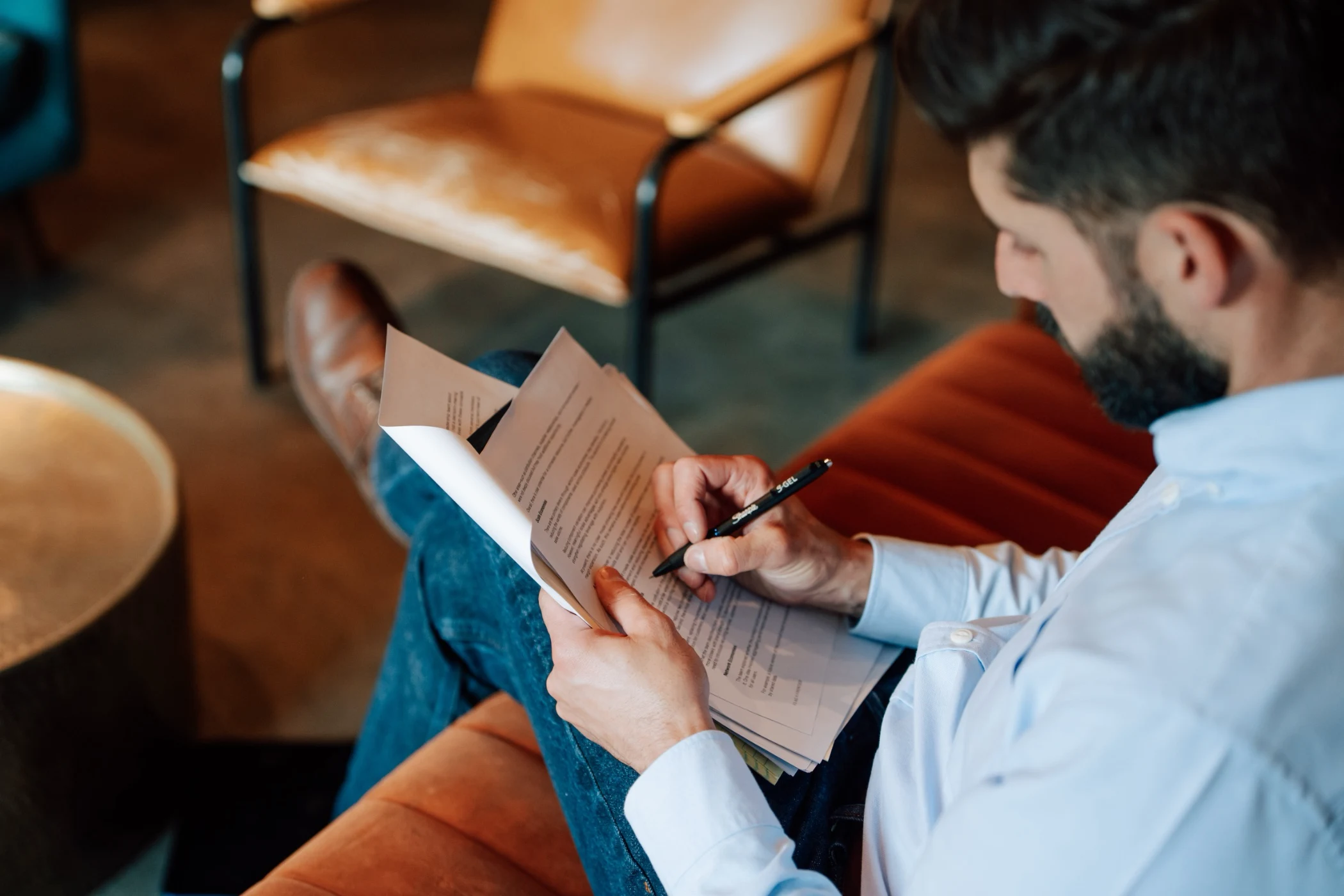 Man with a beard sitting on an orange couch, writing on papers with a pen.