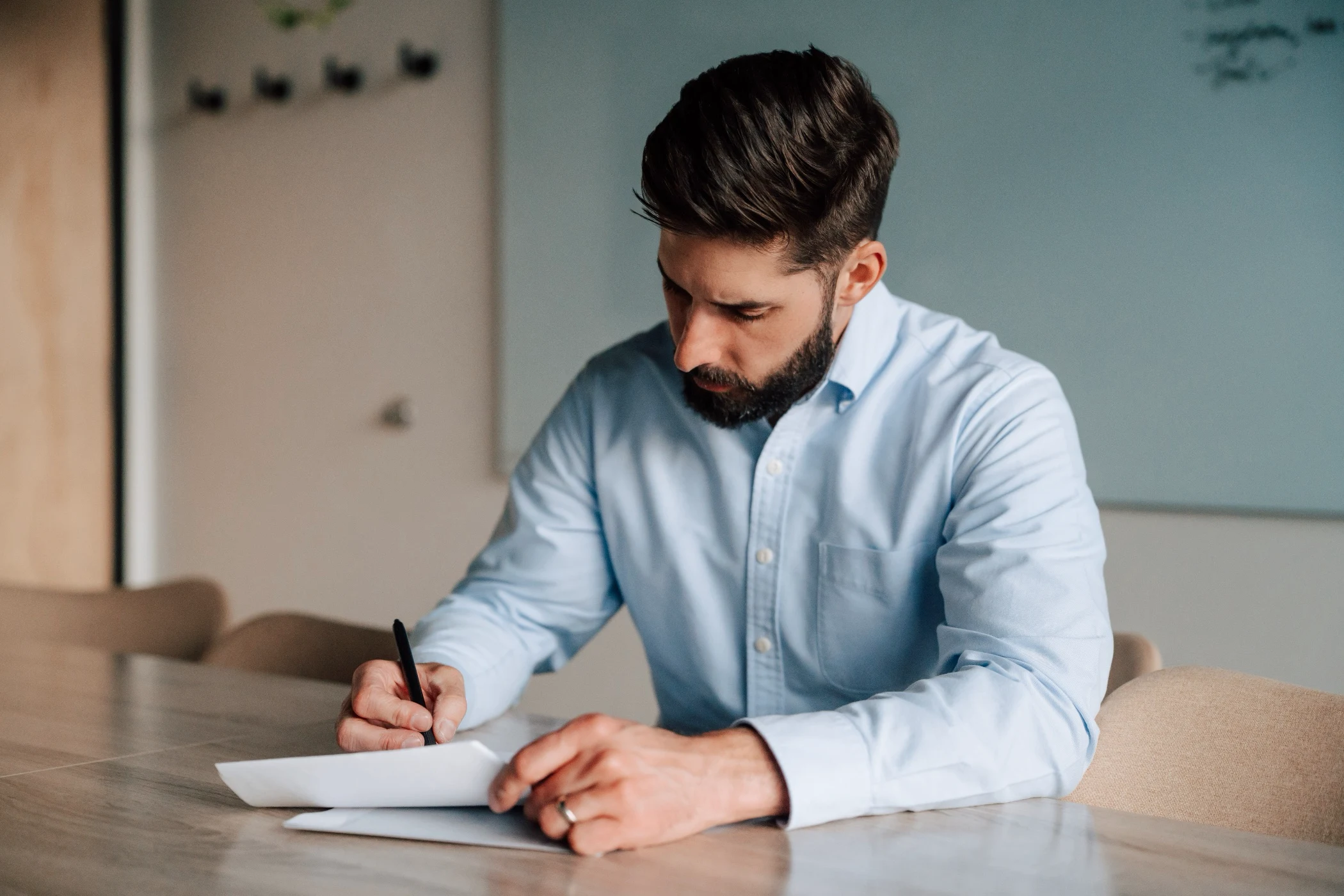 Man with dark hair and beard in a light blue shirt sitting at a table, writing on a document with a pen.