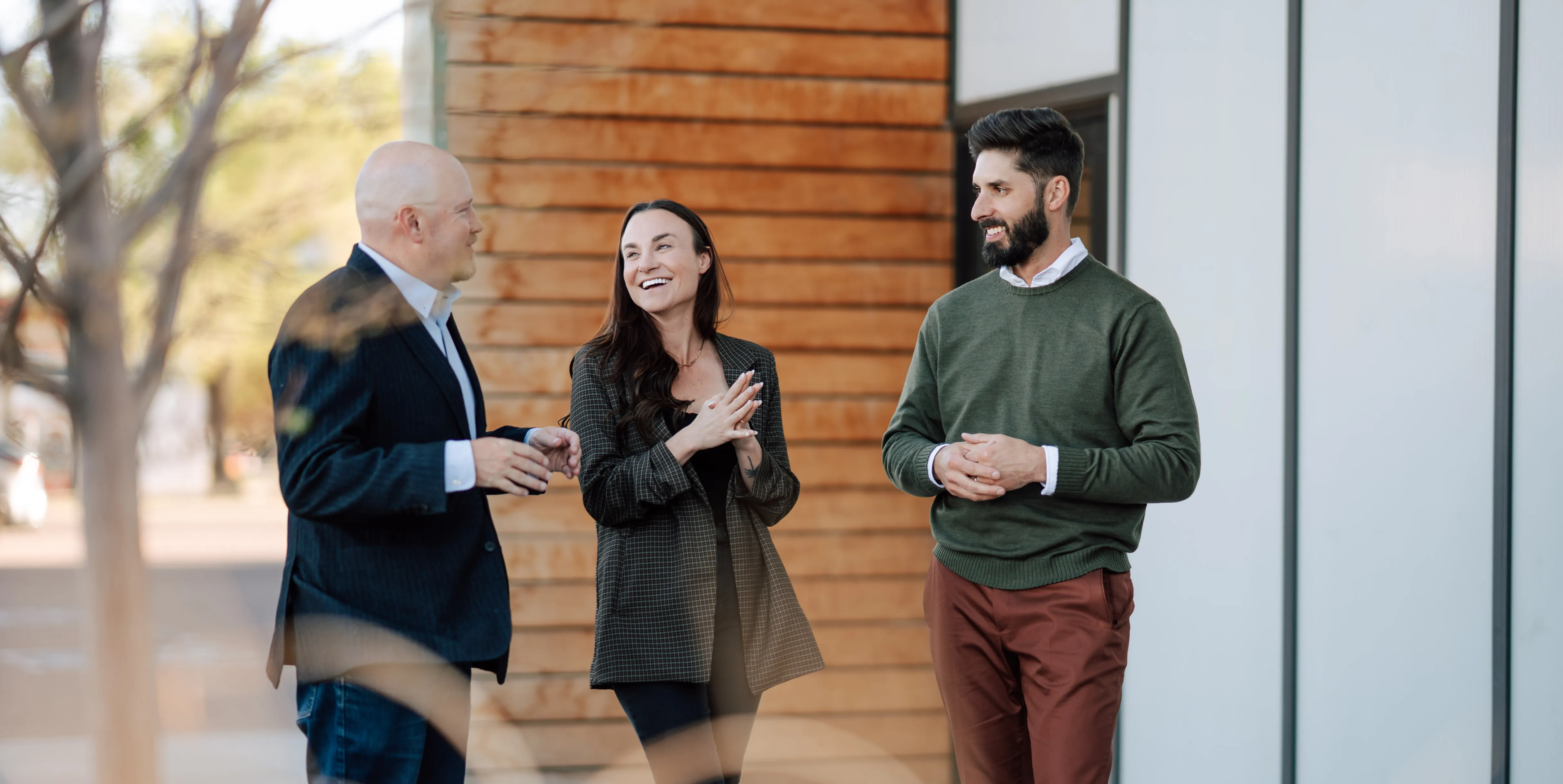 Three casually dressed people standing outdoors smiling and talking near a wooden building.