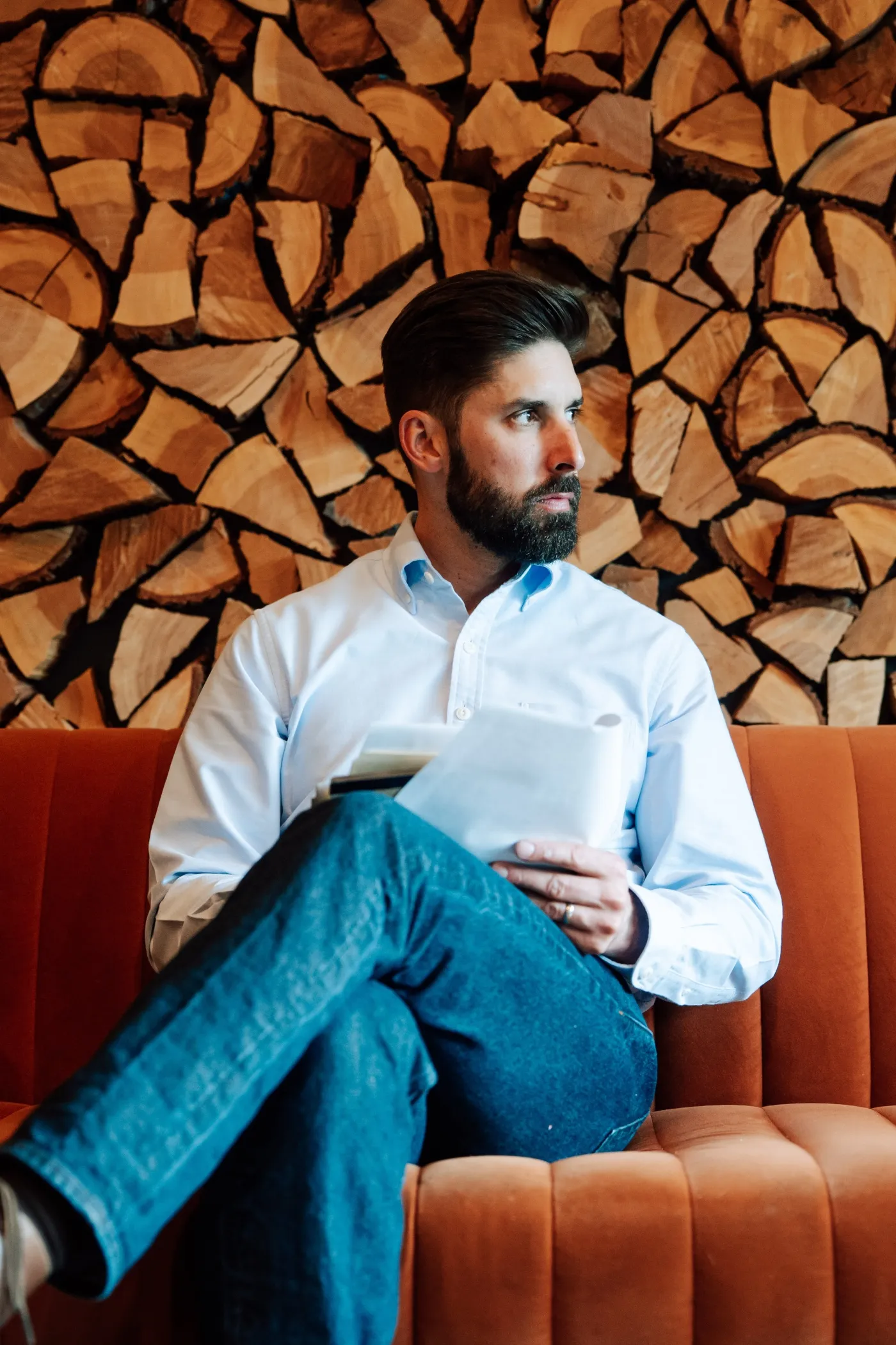 Bearded man in blue shirt sitting on orange couch holding papers, wood stack wall behind him.