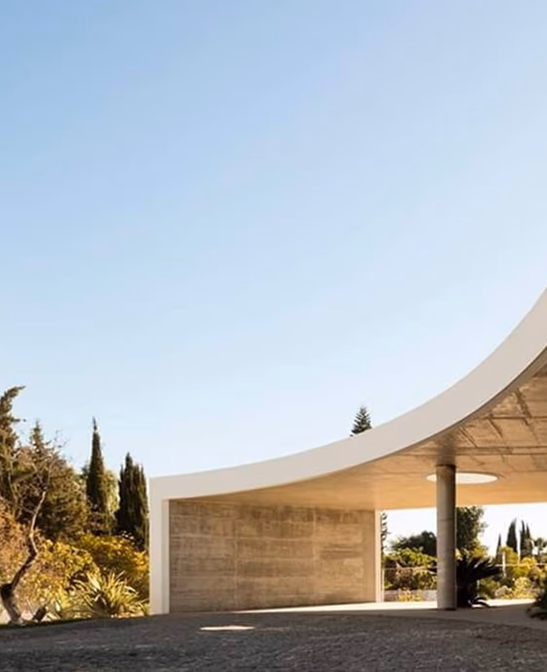 Modern curved concrete structure with open pillars surrounded by trees and bushes under a clear blue sky.