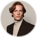 Young man with light brown hair wearing a beige turtleneck and dark brown blazer, facing forward.