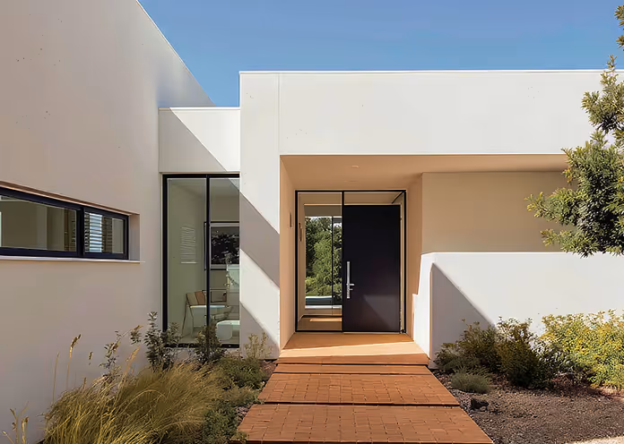 Modern white house entrance with a black door, floor-to-ceiling glass panel, and a walkway with plants on both sides.