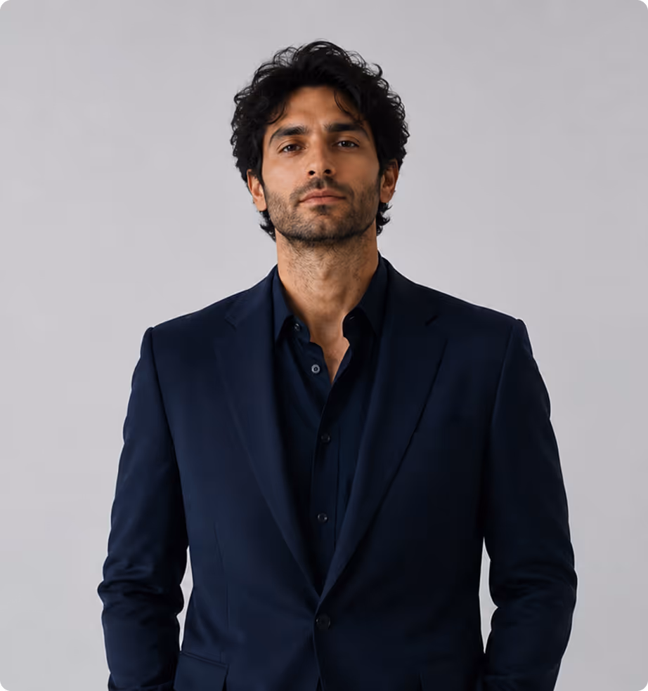 Man with dark curly hair wearing a navy blue suit jacket and shirt against a plain gray background.