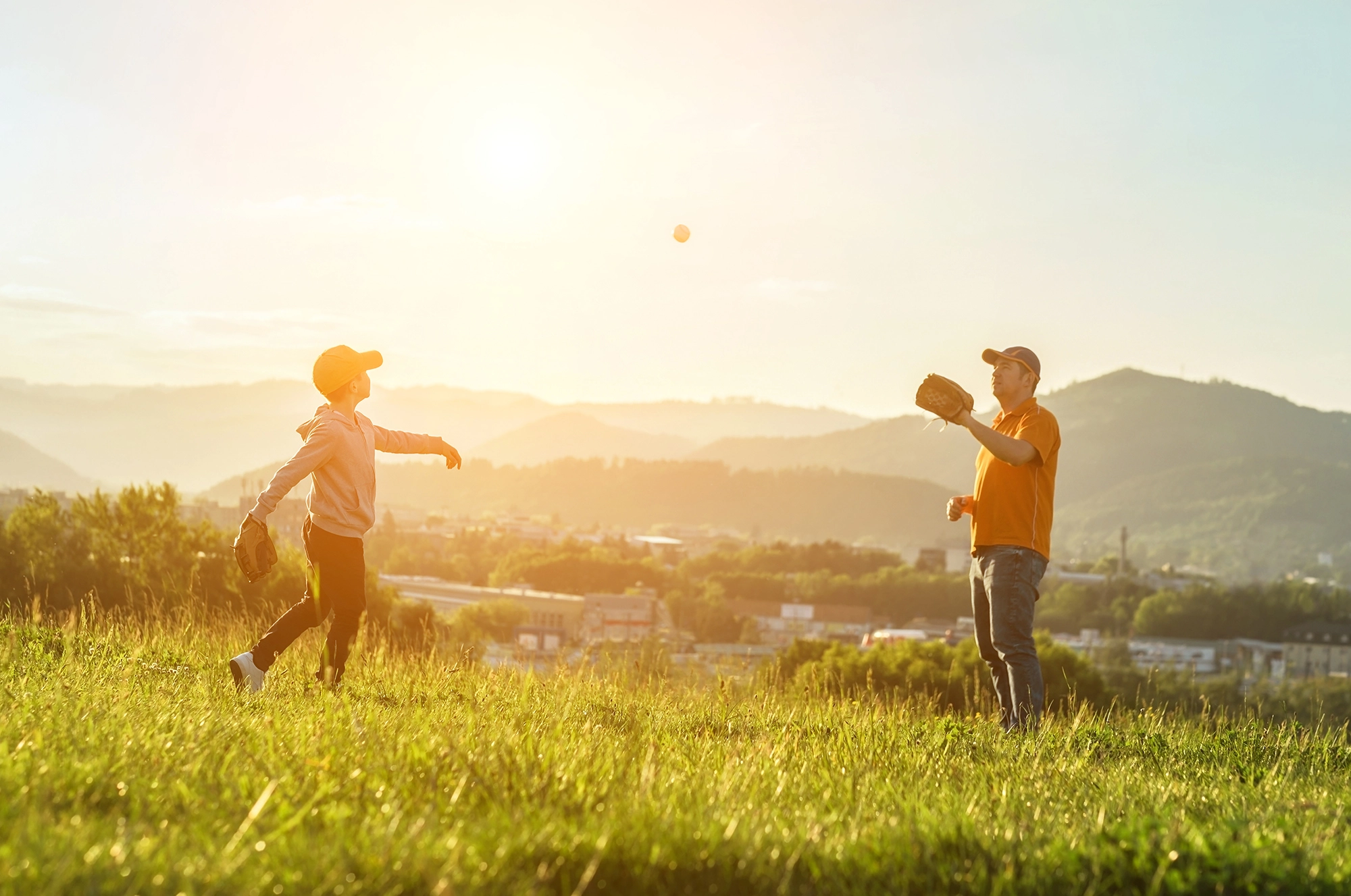 An image of a father and son bonding by playing catch on a beautiful sunny day. The photo captures the moment the boy releases the ball, his arm outstretched as the baseball flies through the hazy sky. The boy's father readies his glove to catch it.