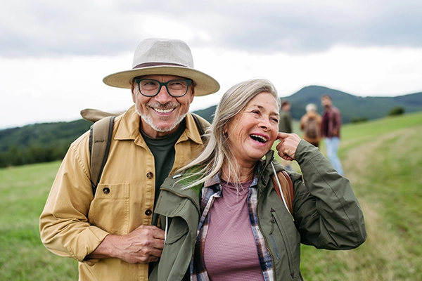 An older man wearing a wide brimmed hat smiles playfully as he embraces his wife's arm. The woman is chuckling and brushing her graying hair from her cheek. They are enjoying a day out hiking with their children and grandchildren. Their joyful faces bare the wrinkles of years filled will love and laughter.