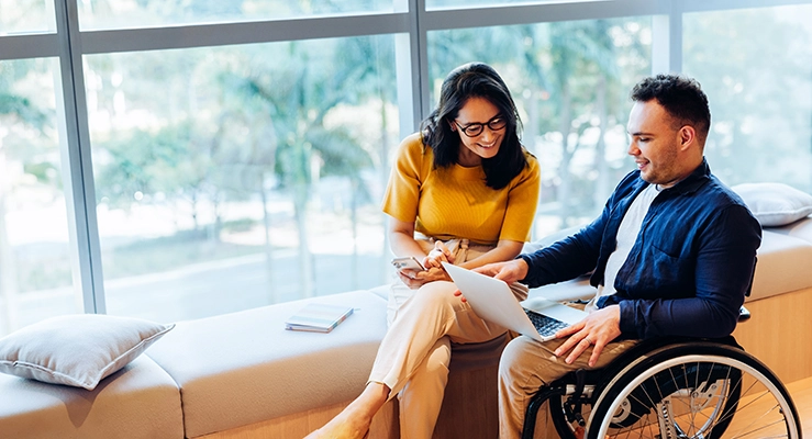 A woman with glasses and a man in a wheelchair look at a laptop together. They are smiling as they talk and navigate their goals with confidence and ease.