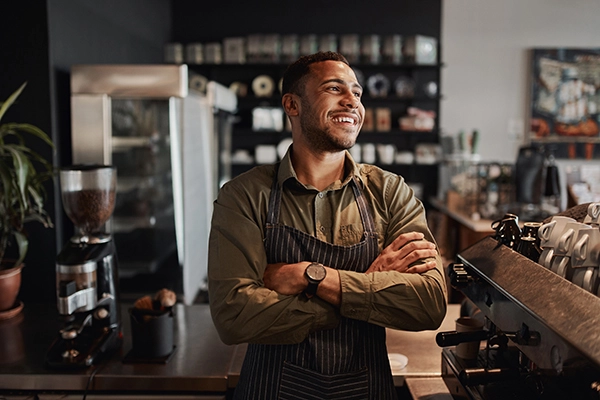 This small business owner is a young and confident man of color. He smiles joyfully, as if taking a moment to soak in his accomplishments, in the coffee shop he owns and operates.