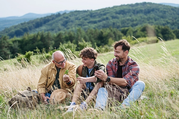 Sitting in the wild grass on a sprawling hillside are a grandfather, a son, and a grandson. The eldest appears to be passing wisdom down to the other two.