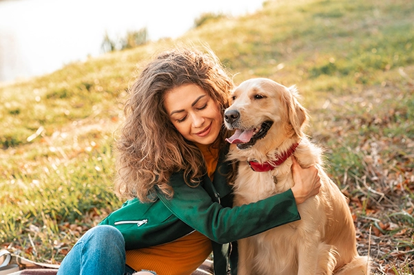 A woman with long, wavy brown hair hugs a happy golden retriever on a sunny day in the park.
