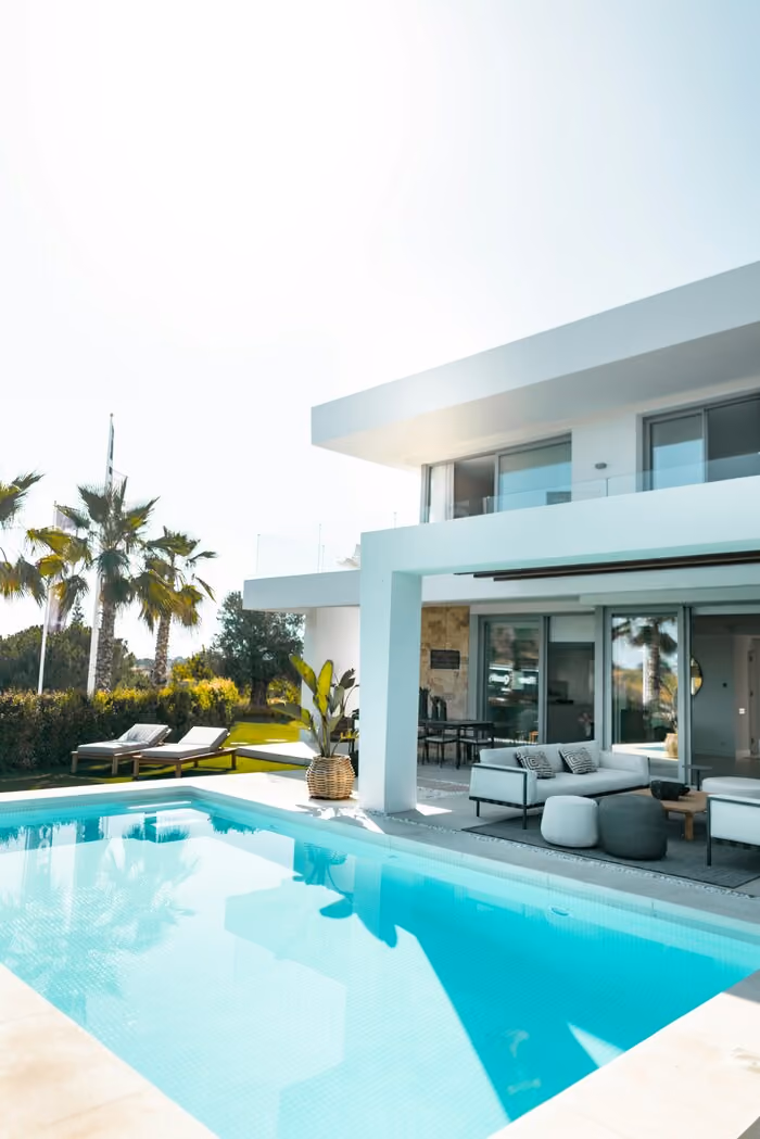 Modern white house with a patio, outdoor seating, palm trees, and a clear blue swimming pool.