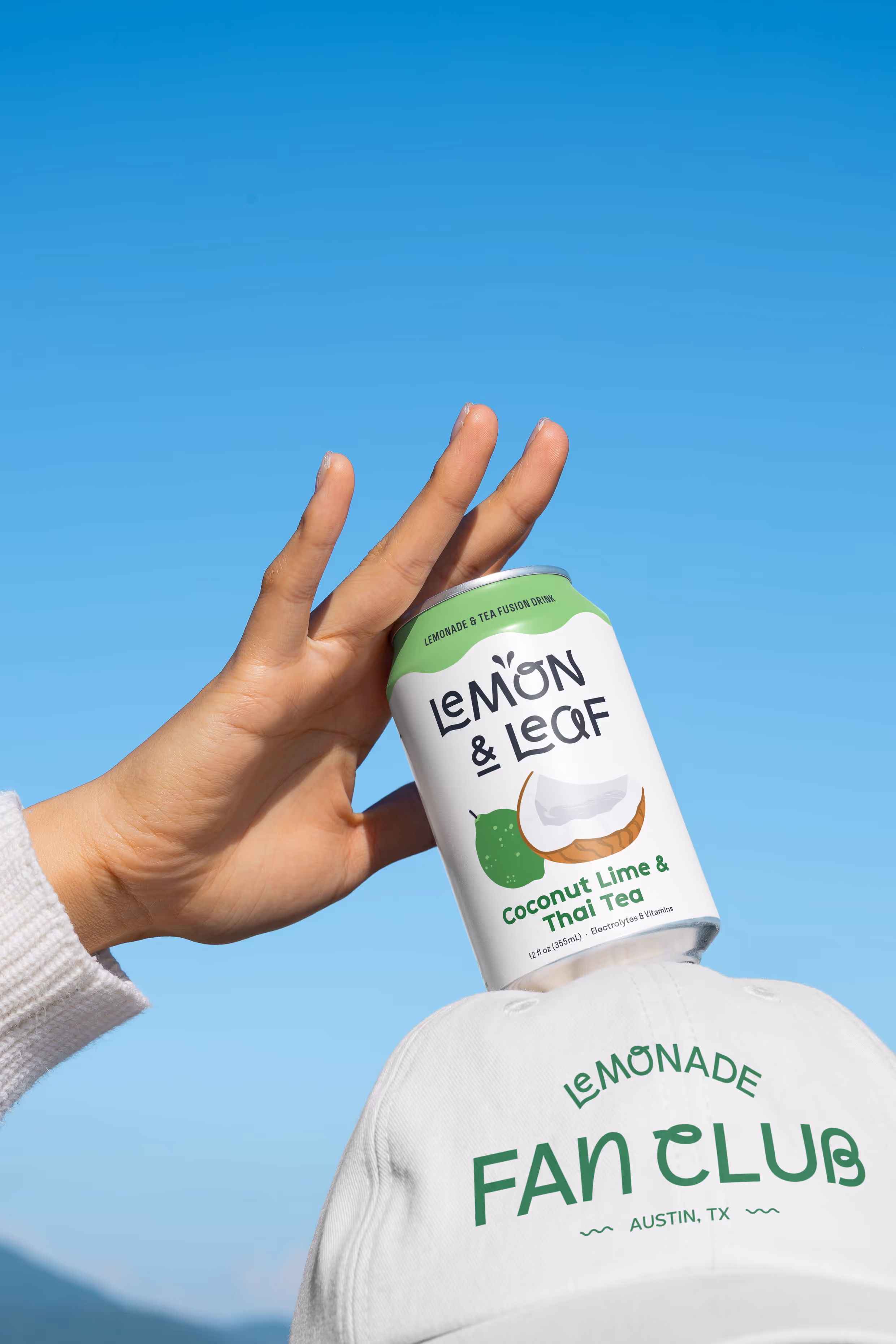 A hand holding a Lemon & Leaf Coconut Lime and Thai Tea can against a blue sky, above a white "Lemonade Fan Club, Austin TX" cap.