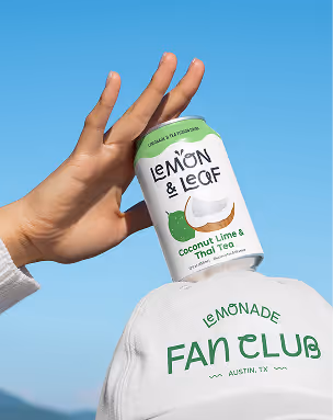 A hand holding a Lemon & Leaf Coconut Lime and Thai Tea can against a blue sky, above a white "Lemonade Fan Club, Austin TX" cap.
