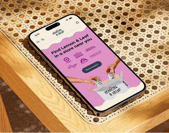 An iPhone on a woven rattan table showing the Lemon & Leaf store locator, its pink screen reading "Find Lemon & Leaf in a store near you" above a row of retailer logos.