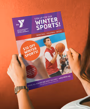 Hands holding a purple and orange YMCA winter sports flyer advertising $10 off basketball, volleyball and soccer, with a photo of a young player holding a basketball.