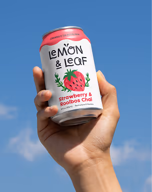 A hand raising a Lemon & Leaf Strawberry & Rooibos Chai can against a clear blue sky.