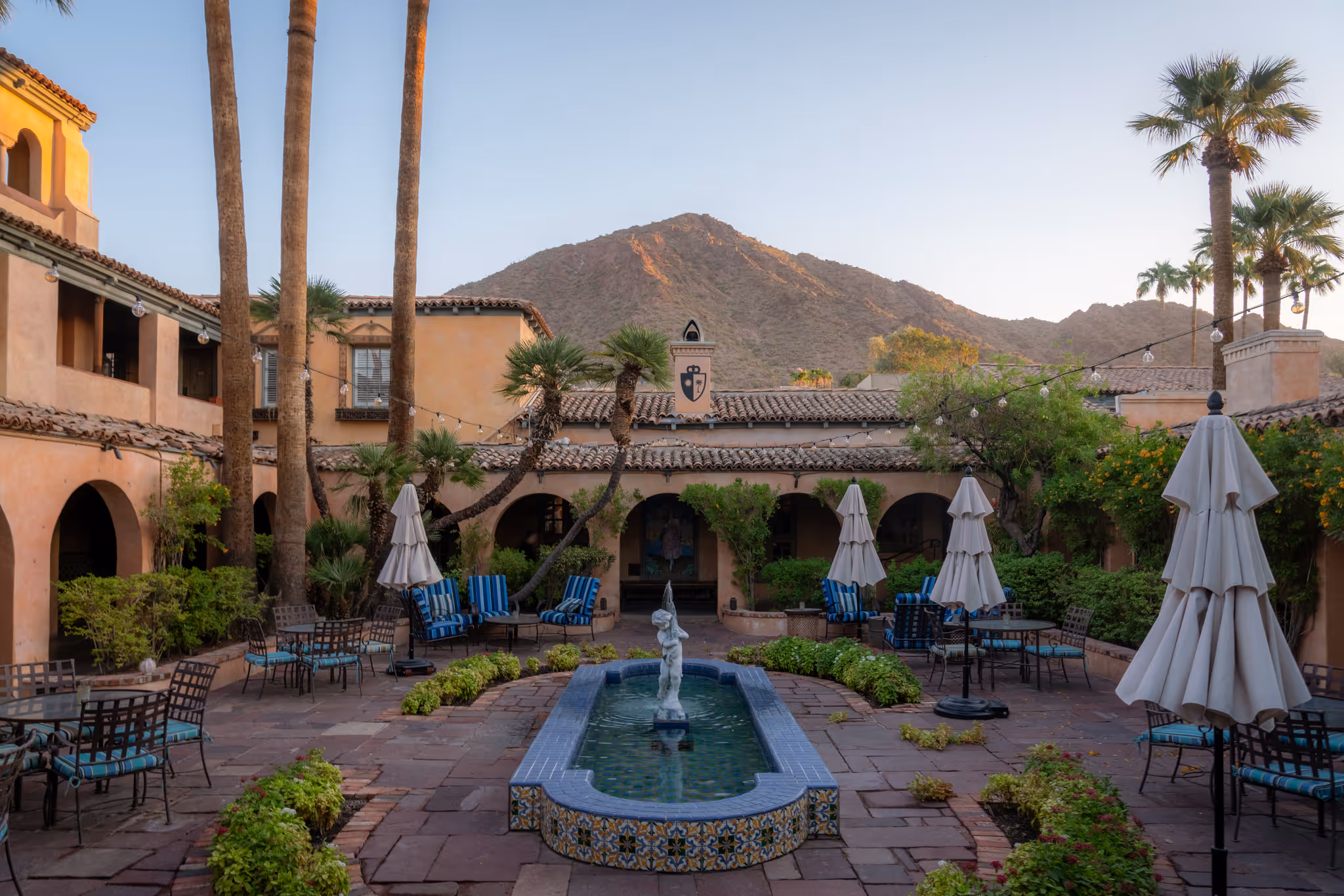 Sunny courtyard with fountain, palm trees, chairs, umbrellas, and mountain backdrop at sunset.