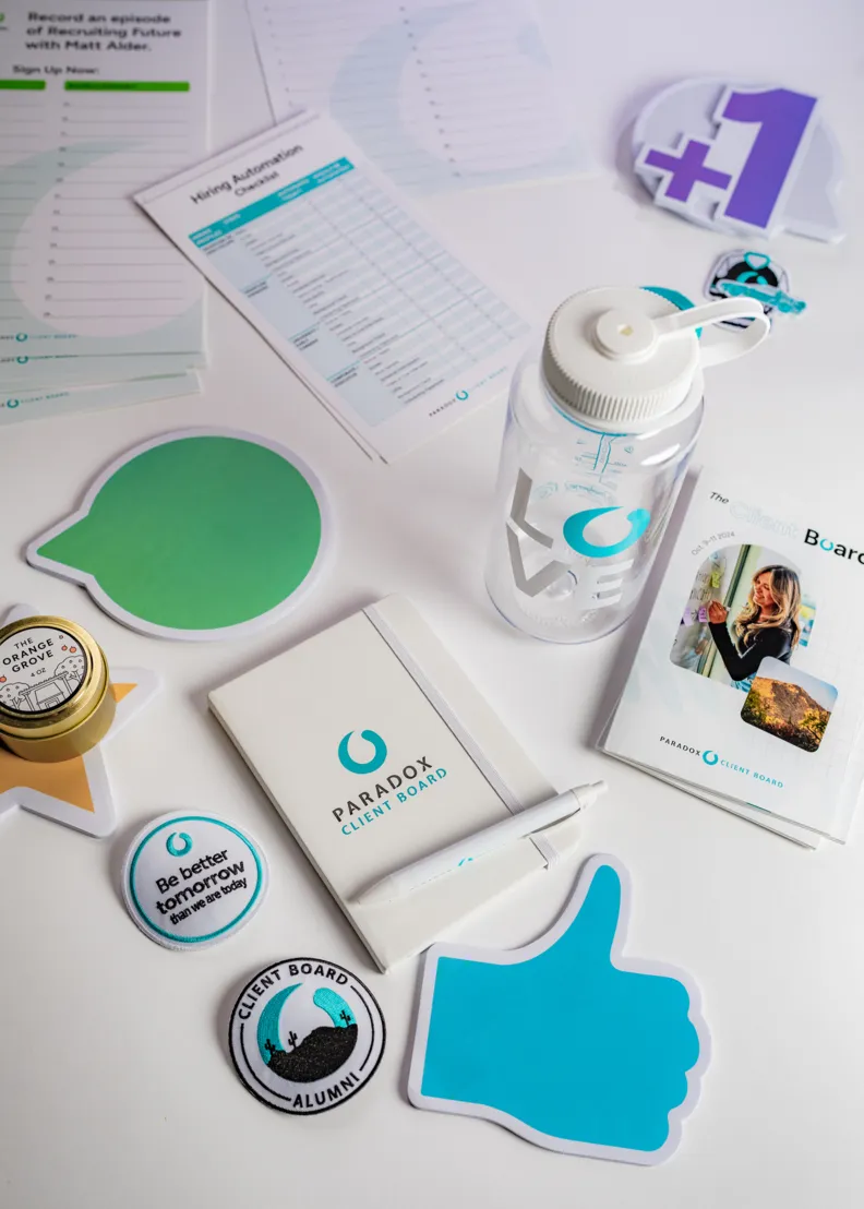Paradox Client Board branded items including notebook, water bottle, stickers, and pins on a white surface.