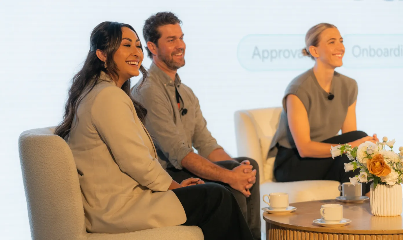 Three people seated in chairs smiling during a panel discussion with a table holding cups and flowers.