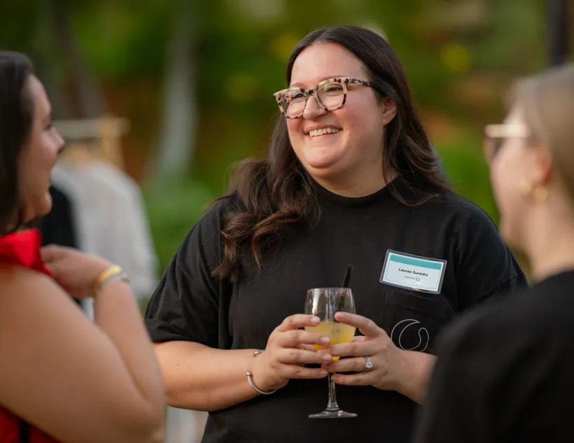 Smiling woman in glasses and black shirt holding a drink talks with two other women outdoors.