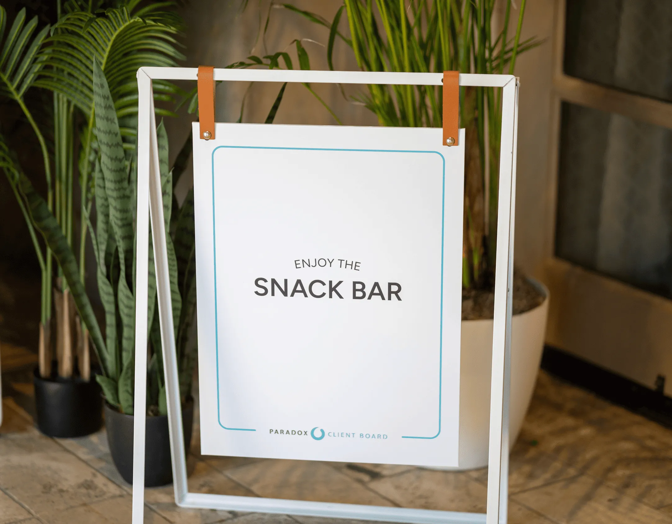 White sign on stand reading 'Enjoy the Snack Bar' with plants in the background.