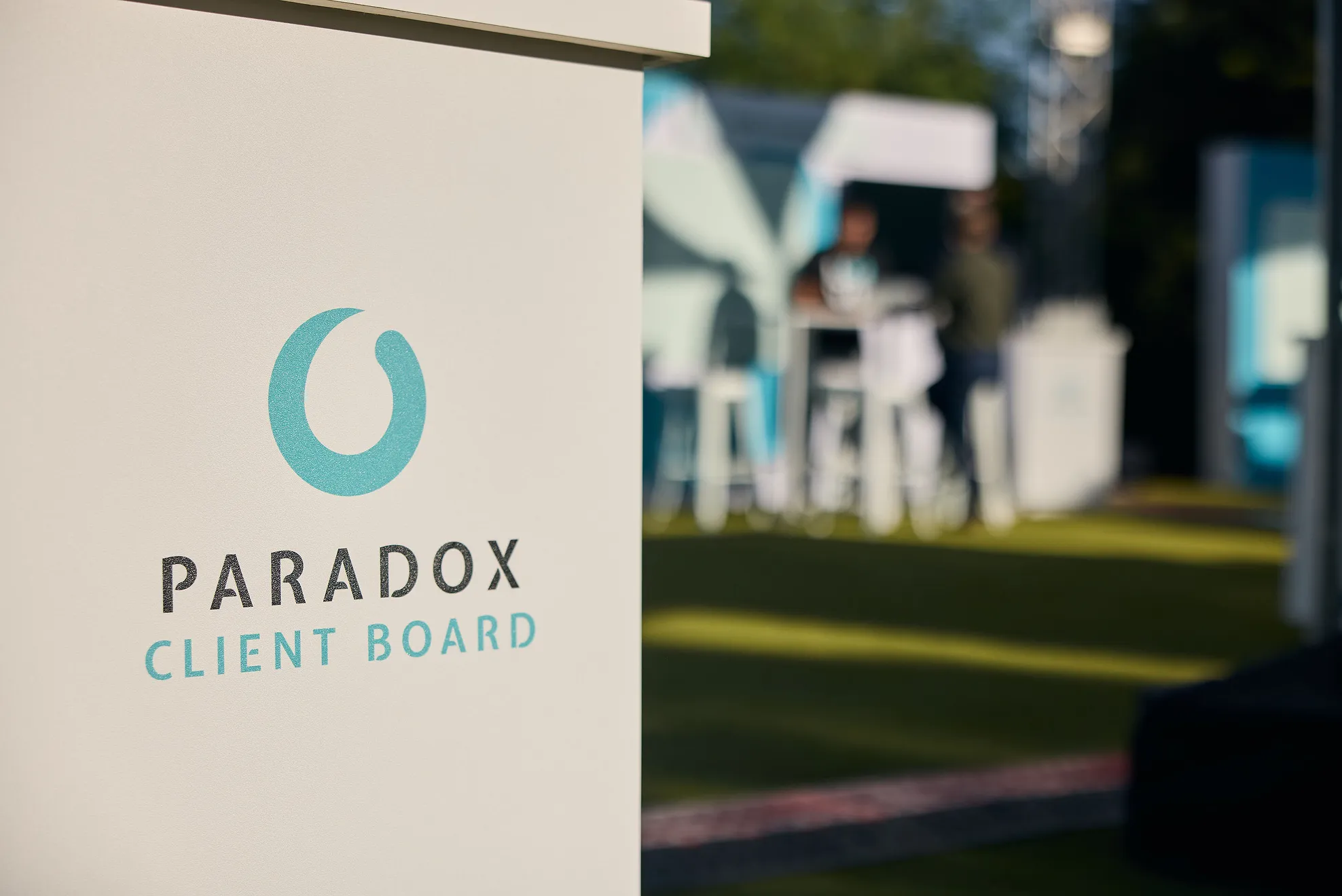 White board with turquoise logo and black text reading 'Paradox Client Board' outdoors with blurred people.