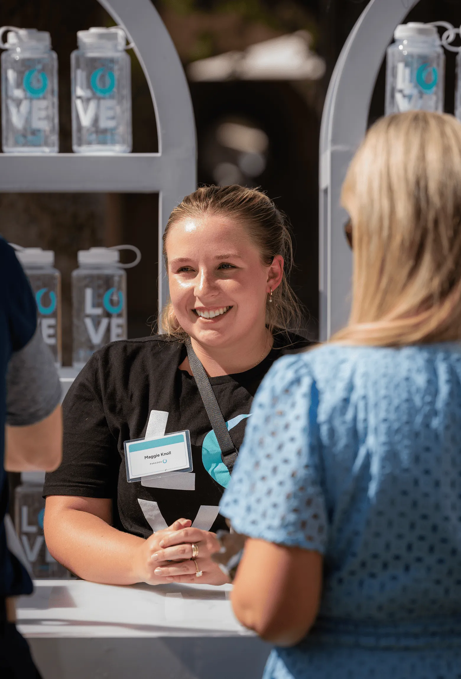 Smiling woman wearing a name tag talks to a person in a blue dress at an outdoor booth.